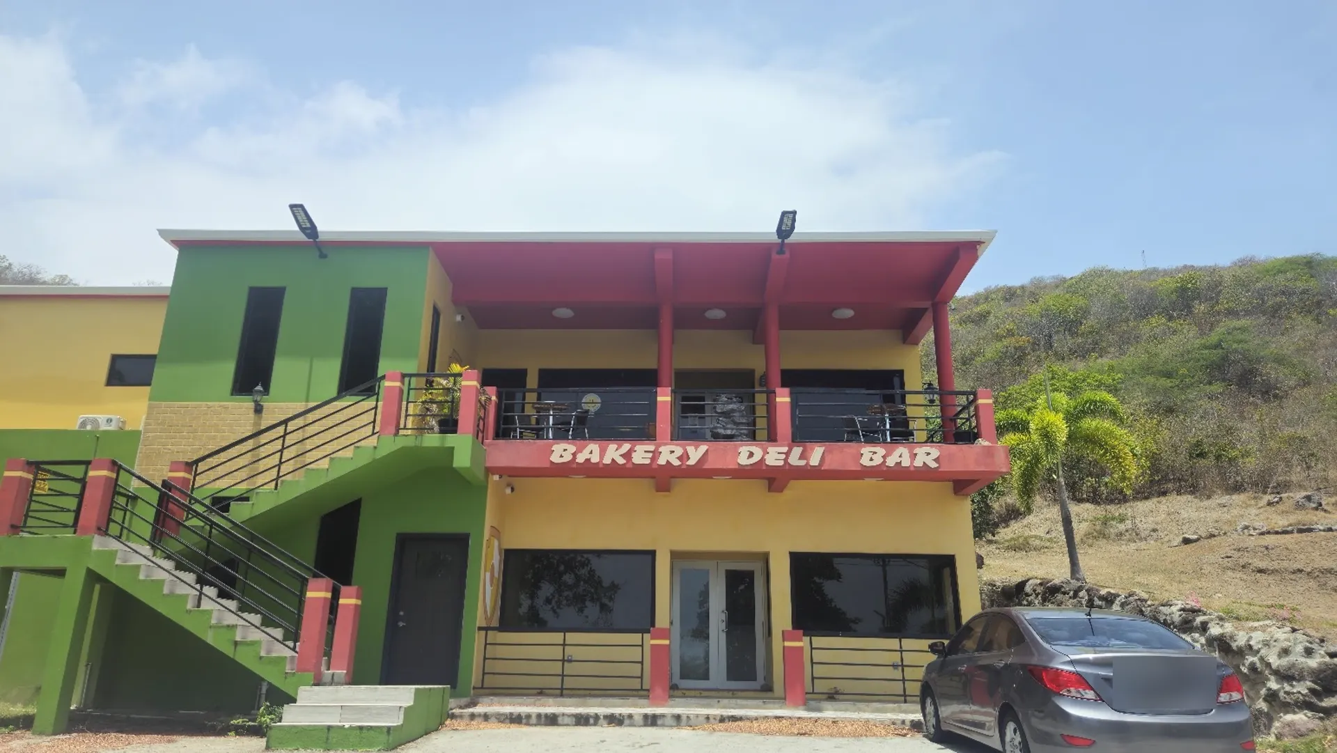 LBW Deli & Bar's yellow and green hillside building overlooking Nevis, with a covered upstairs balcony and the Bakery Deli Bar sign